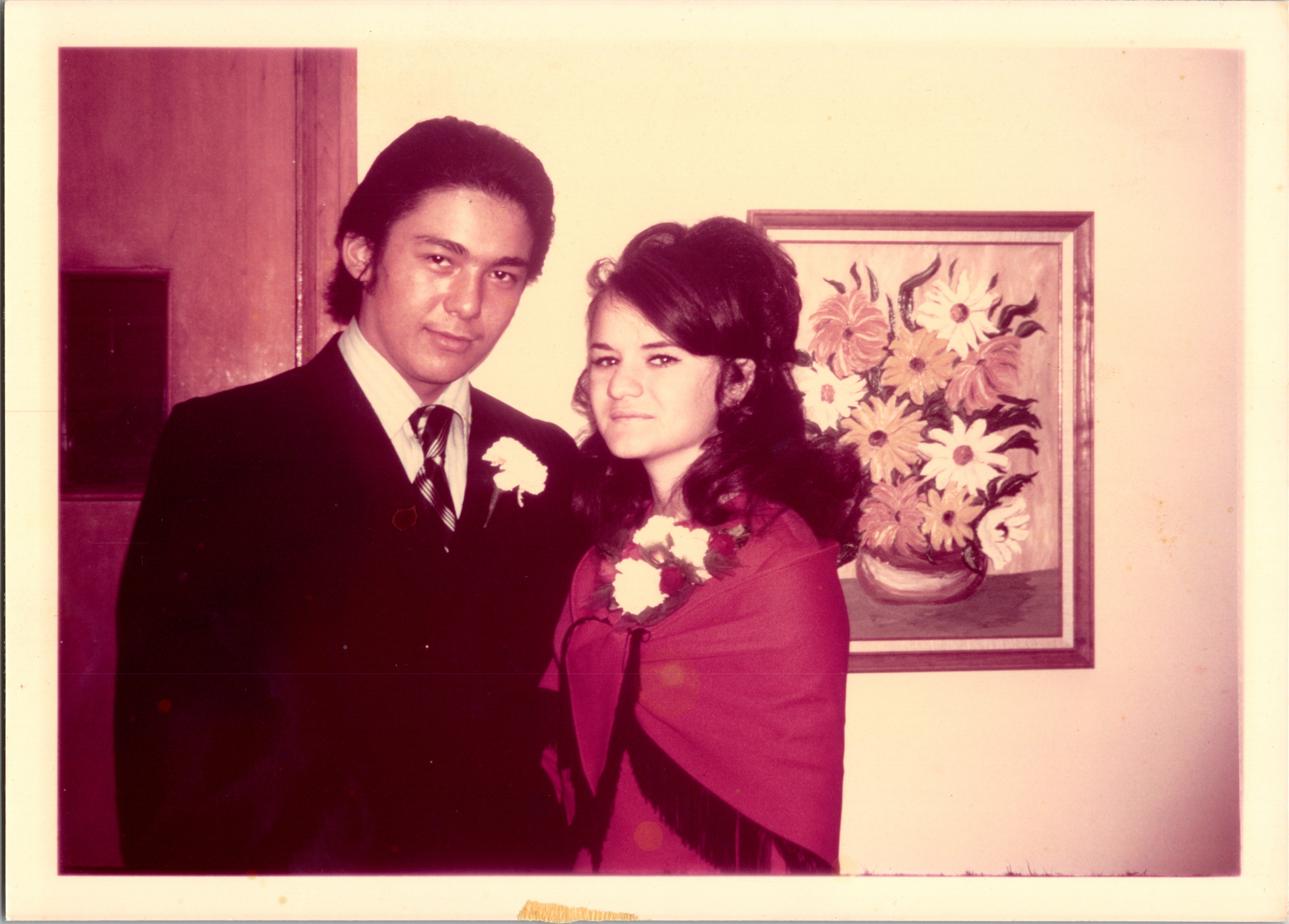 Young couple in formal attire posing indoors, the woman holding a bouquet and wearing a corsage beside the man in a suit.