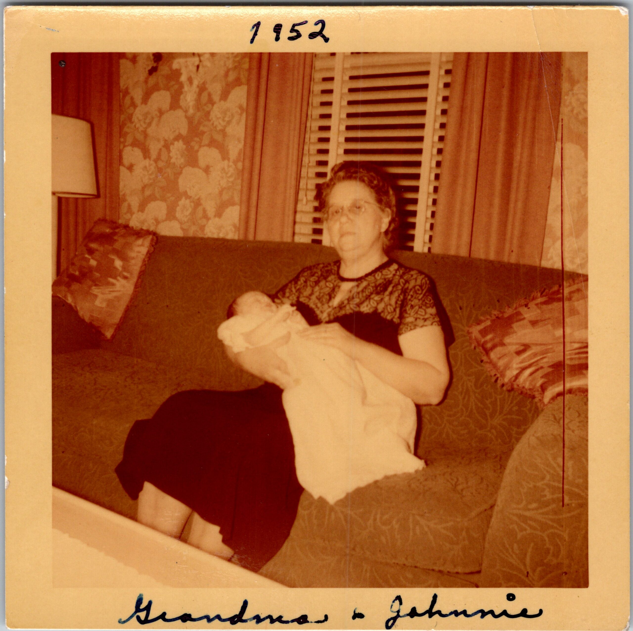 Older woman with glasses holding a small baby on a sofa in a living room with floral wallpaper and blinds behind her.