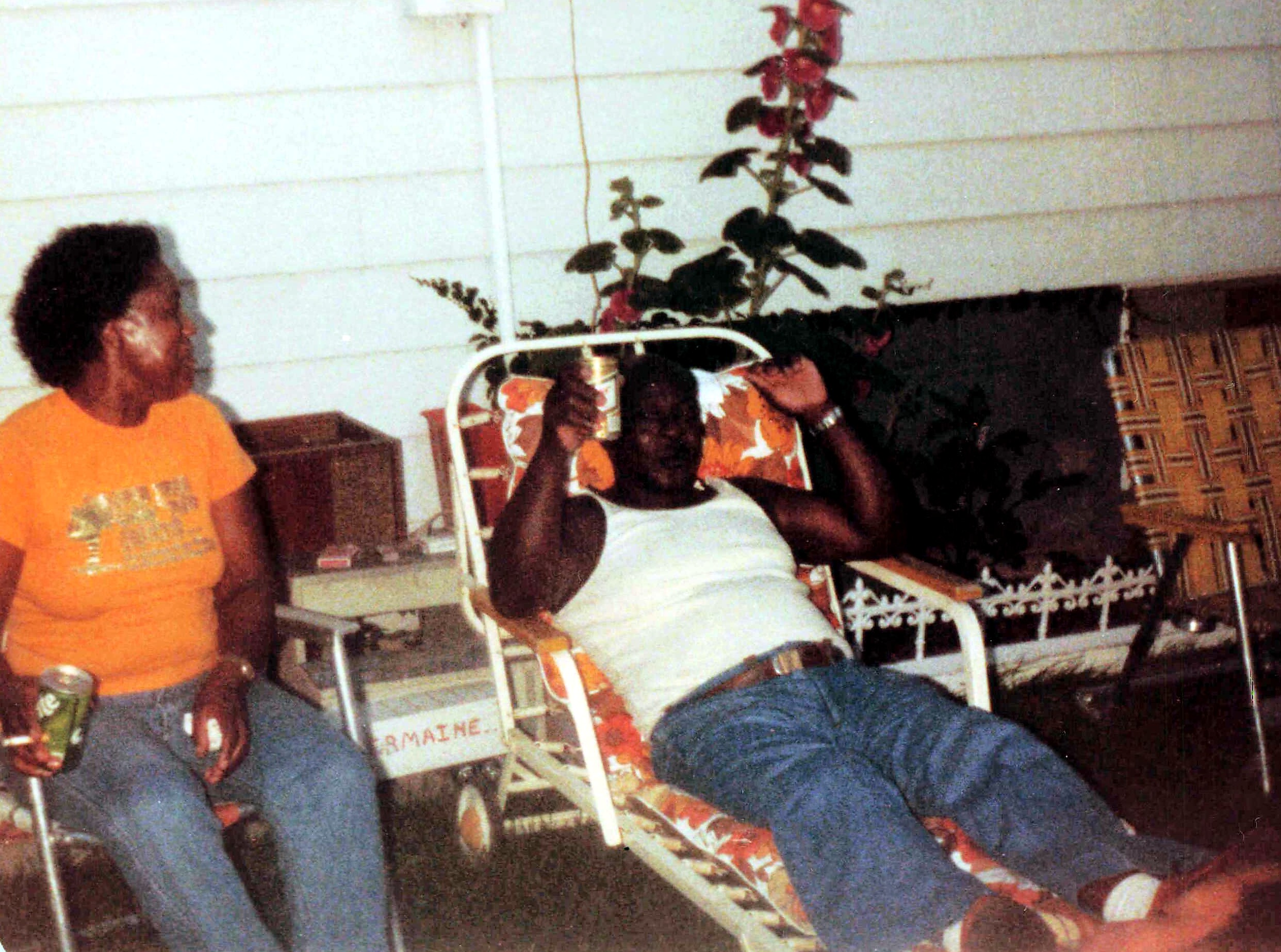 Three people are gathered on a porch: a man in an orange shirt standing, and others seated around a floral chair, with a tall plant in the background.