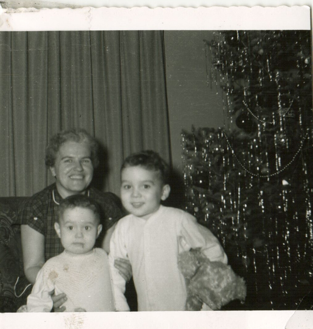 Woman smiling with two young children indoors, posed by a decorated Christmas tree.