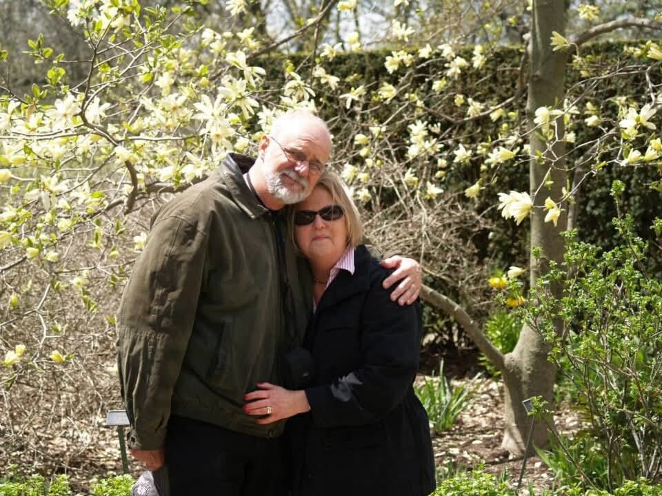Couple embracing in a garden with yellow blossoms on flowering trees behind them.