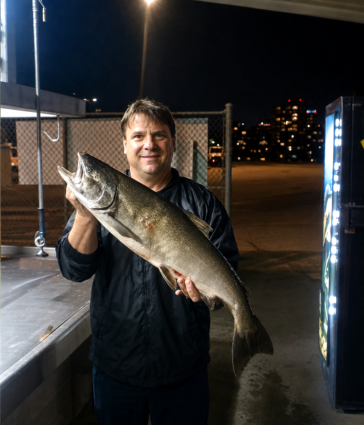 Man in a dark jacket at night holding a large salmon across his chest, outdoors near a fence and vending machine visible to the side.