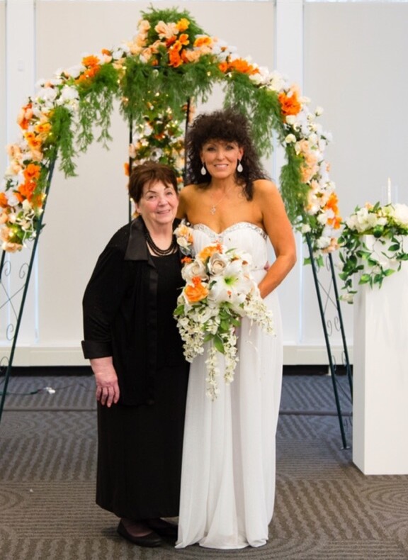Bride in a white strapless gown holding a white and orange bouquet, standing beside an older woman in black under a colorful flower arch.