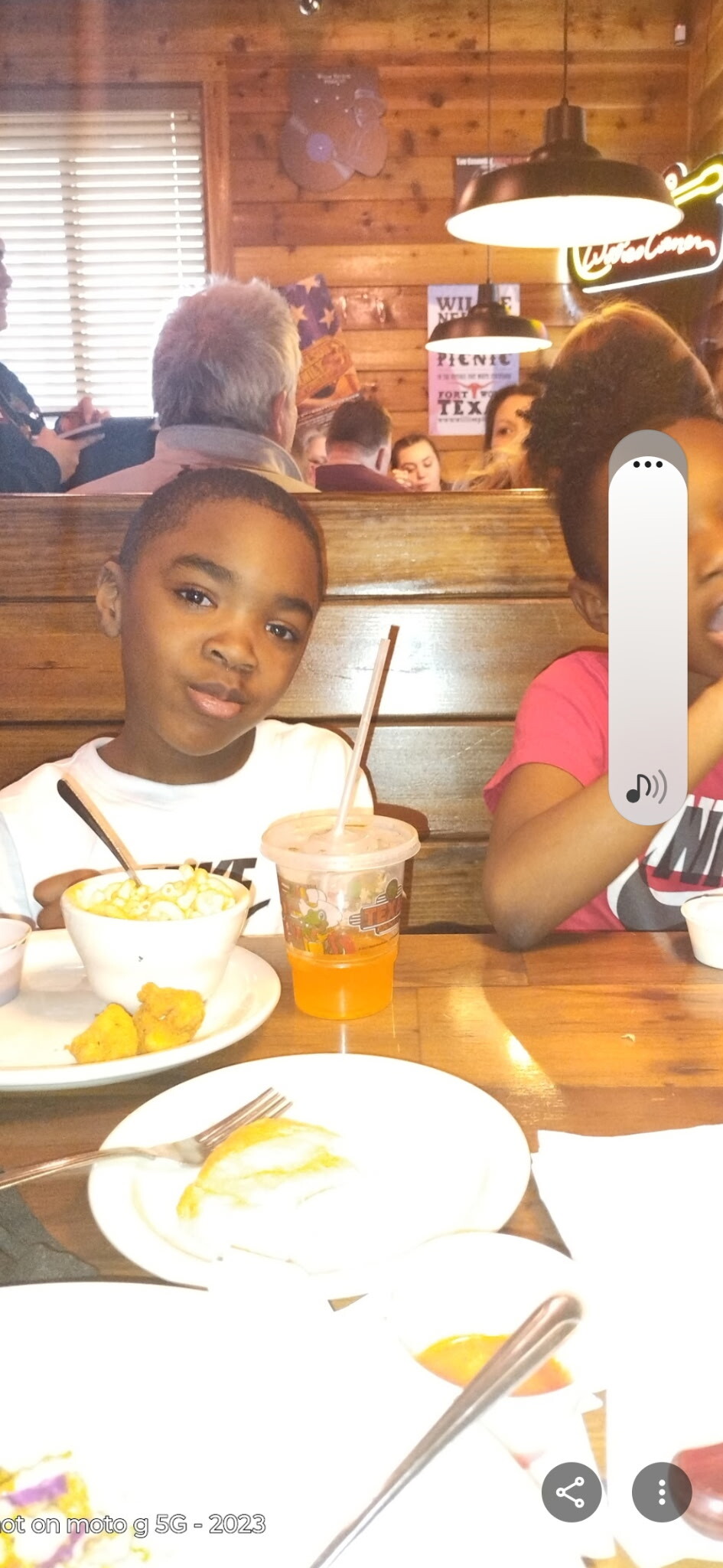 Two kids sit in a wooden booth at a restaurant with drinks and food in front of them, smiling at the camera.