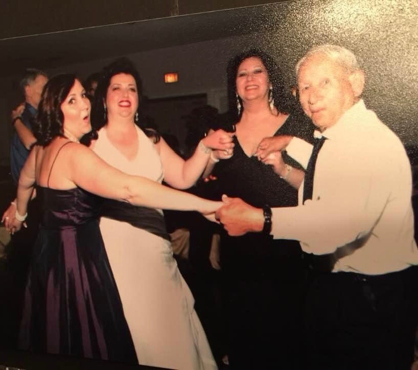 Four adults dancing at a formal event: two women in dresses, one in white, another in purple, with an older man in a white shirt and tie, all smiling and holding hands.