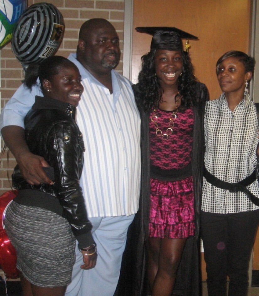 Group of four friends posing indoors at a celebration; a graduate wearing a cap and pink dress stands center with two women and a man nearby.