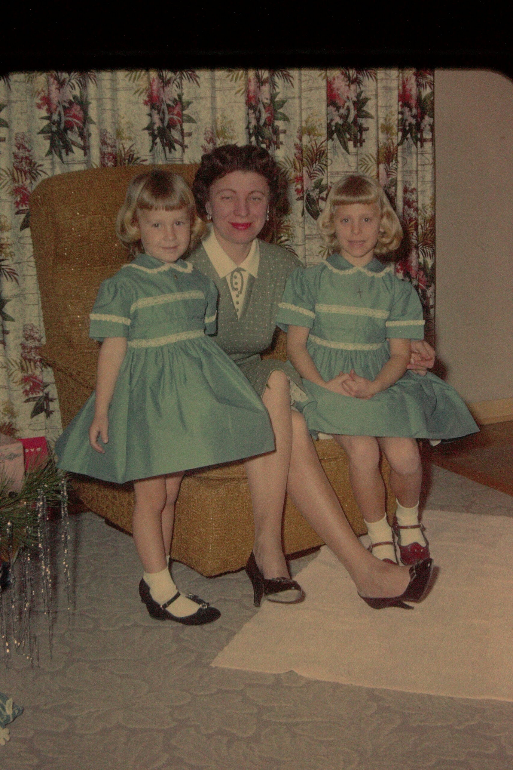 A woman seated between two young girls in matching blue dresses, posing for a family photo on a floral backdrop.