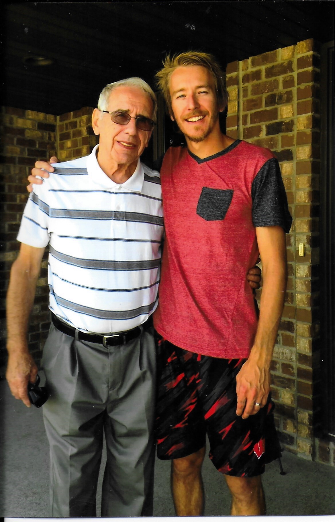 Two men pose with their arms around each other, smiling at the camera outside a brick building.