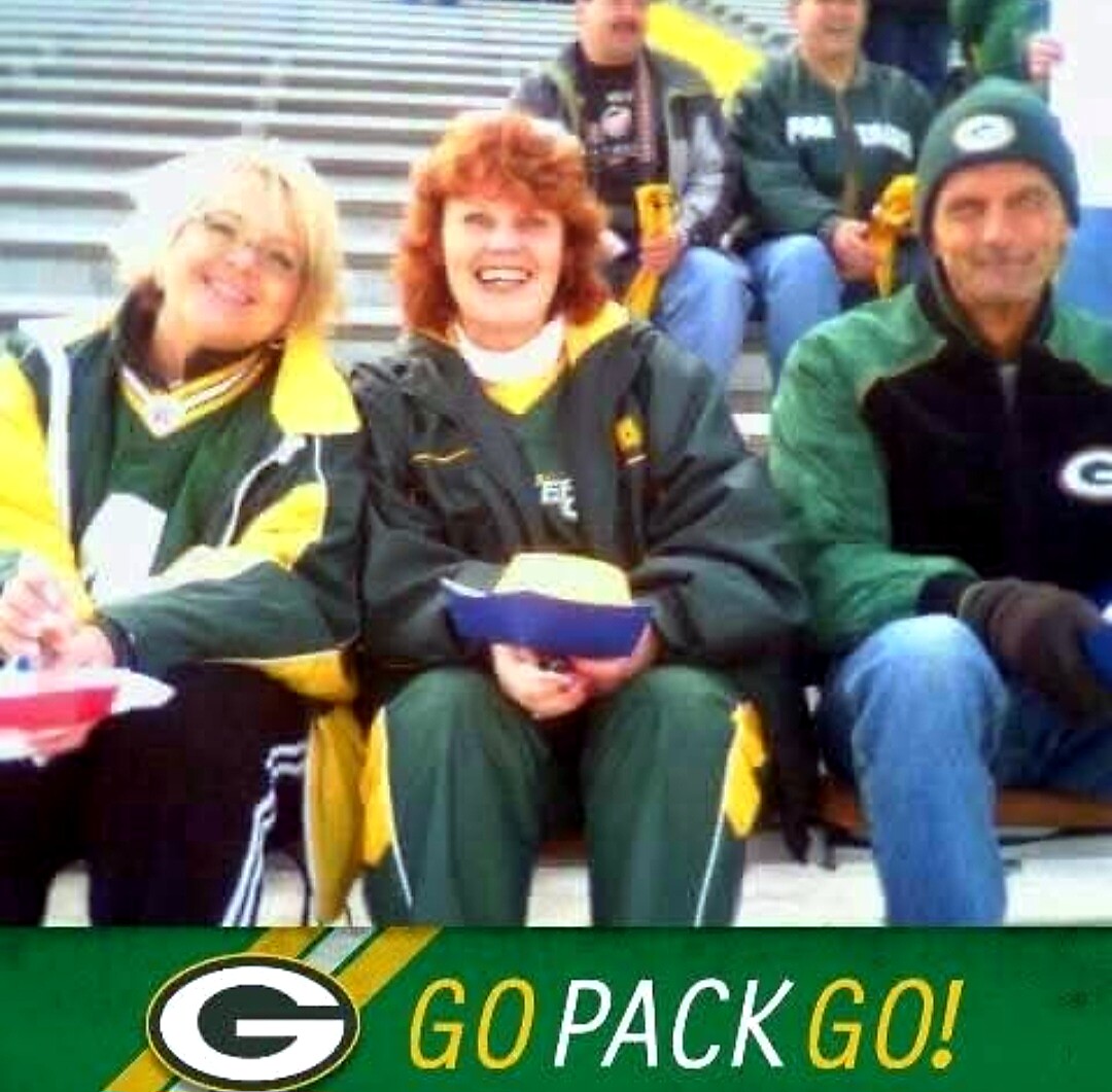 Smiling Packers fans sit on stadium bleachers in green and gold jackets, with a Go Pack Go banner visible at the bottom.