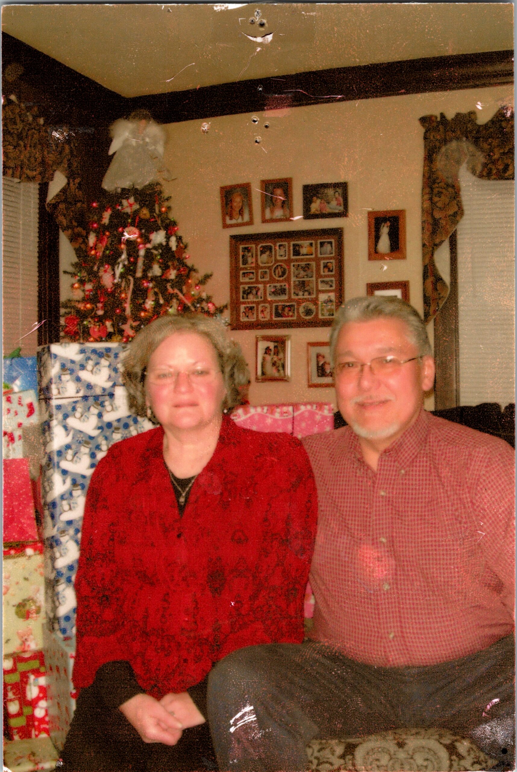 Older couple seated in a living room with a decorated Christmas tree and wrapped gifts behind them.