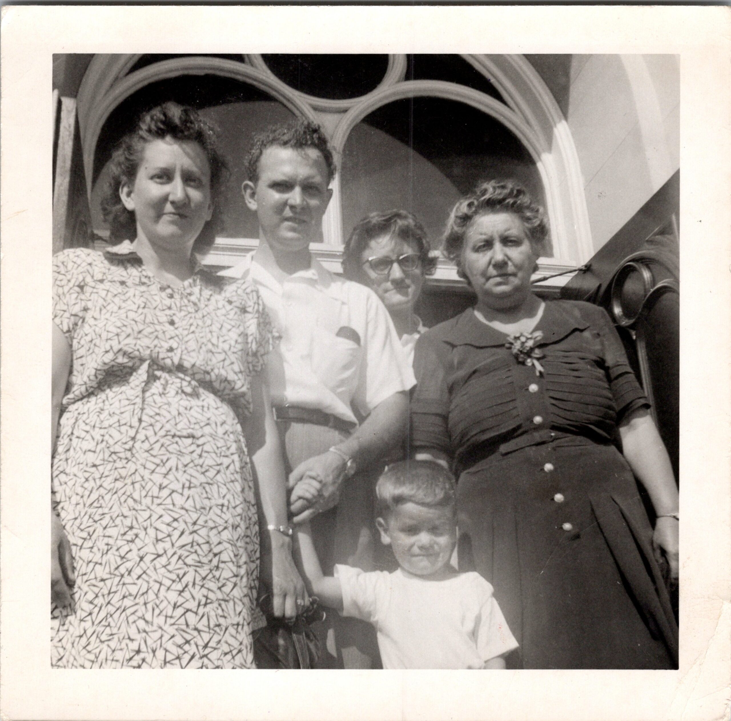 Black-and-white group portrait of four adults and a baby, posing outdoors with curved architectural details in the background; the baby sits in front held by one adult.