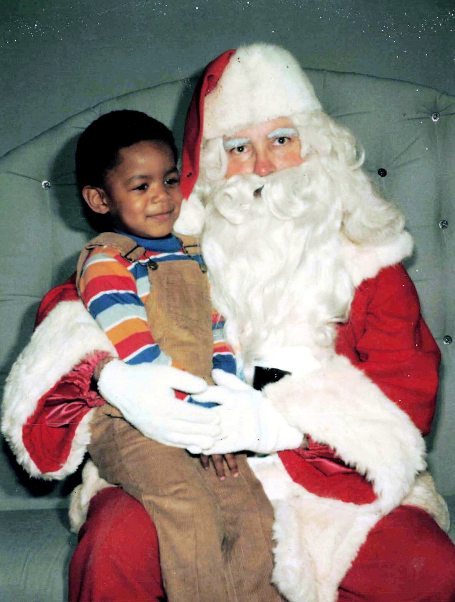 Smiling child sits on Santa's lap, wearing a striped shirt and tan overalls, as Santa in a red suit with a white beard looks on.
