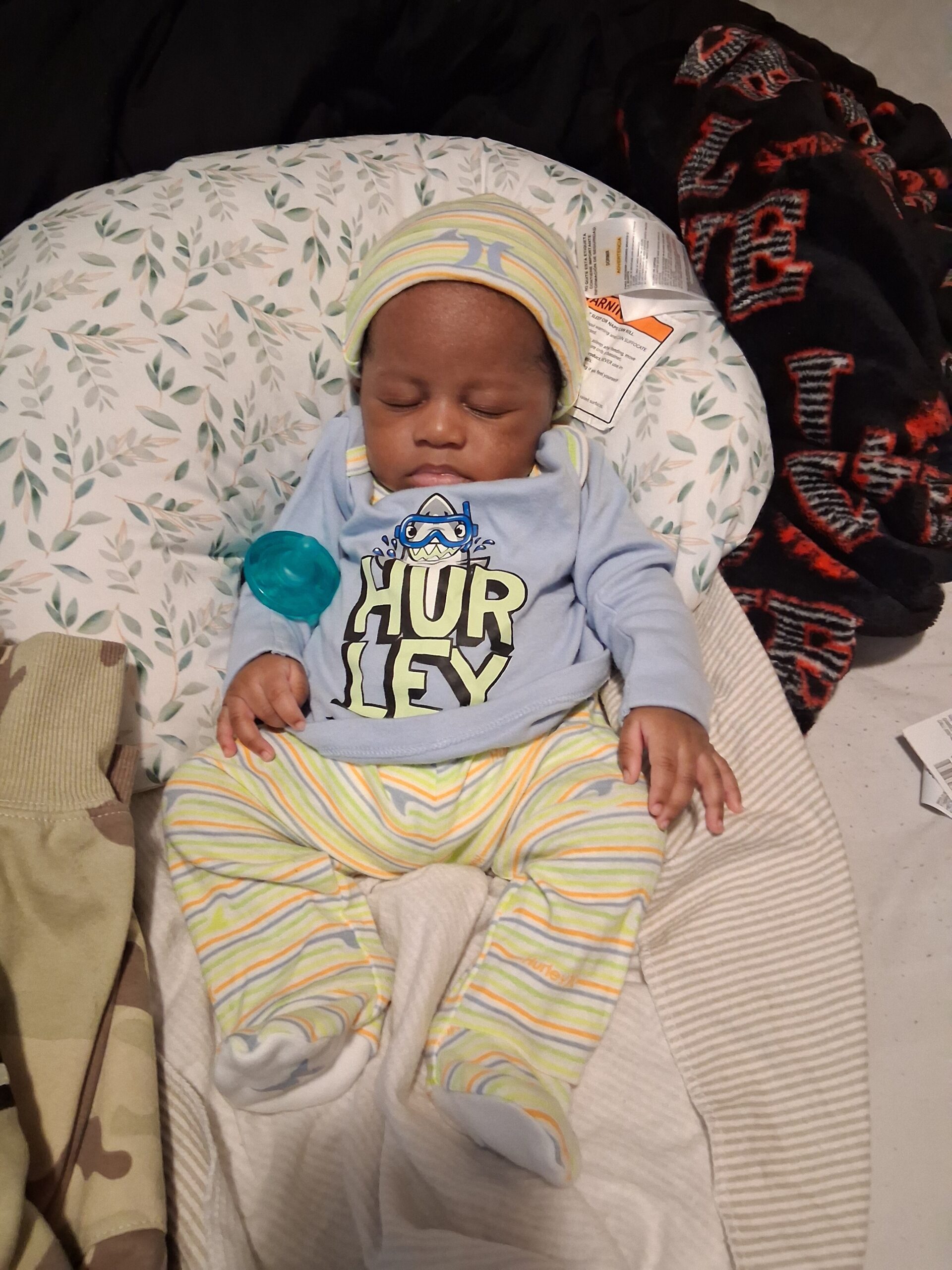 Sleeping baby in pastel striped outfit and hat, resting on a leafy-patterned pillow with a blue pacifier nearby