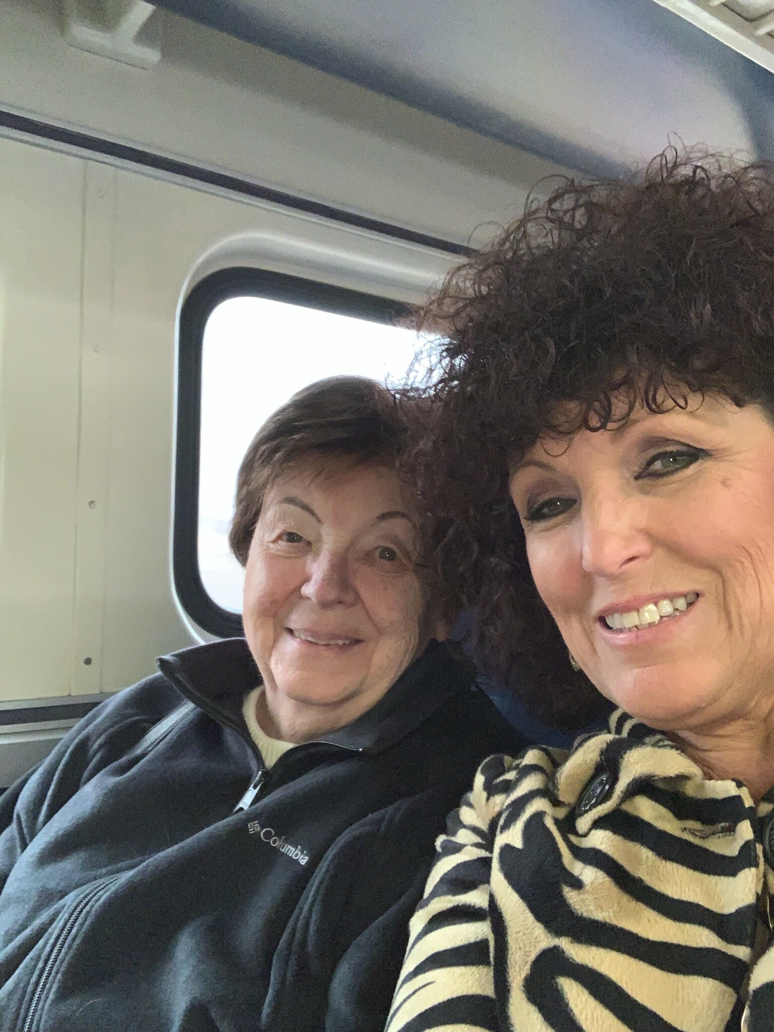 Two women smiling for a selfie inside a vehicle, with a window behind them.