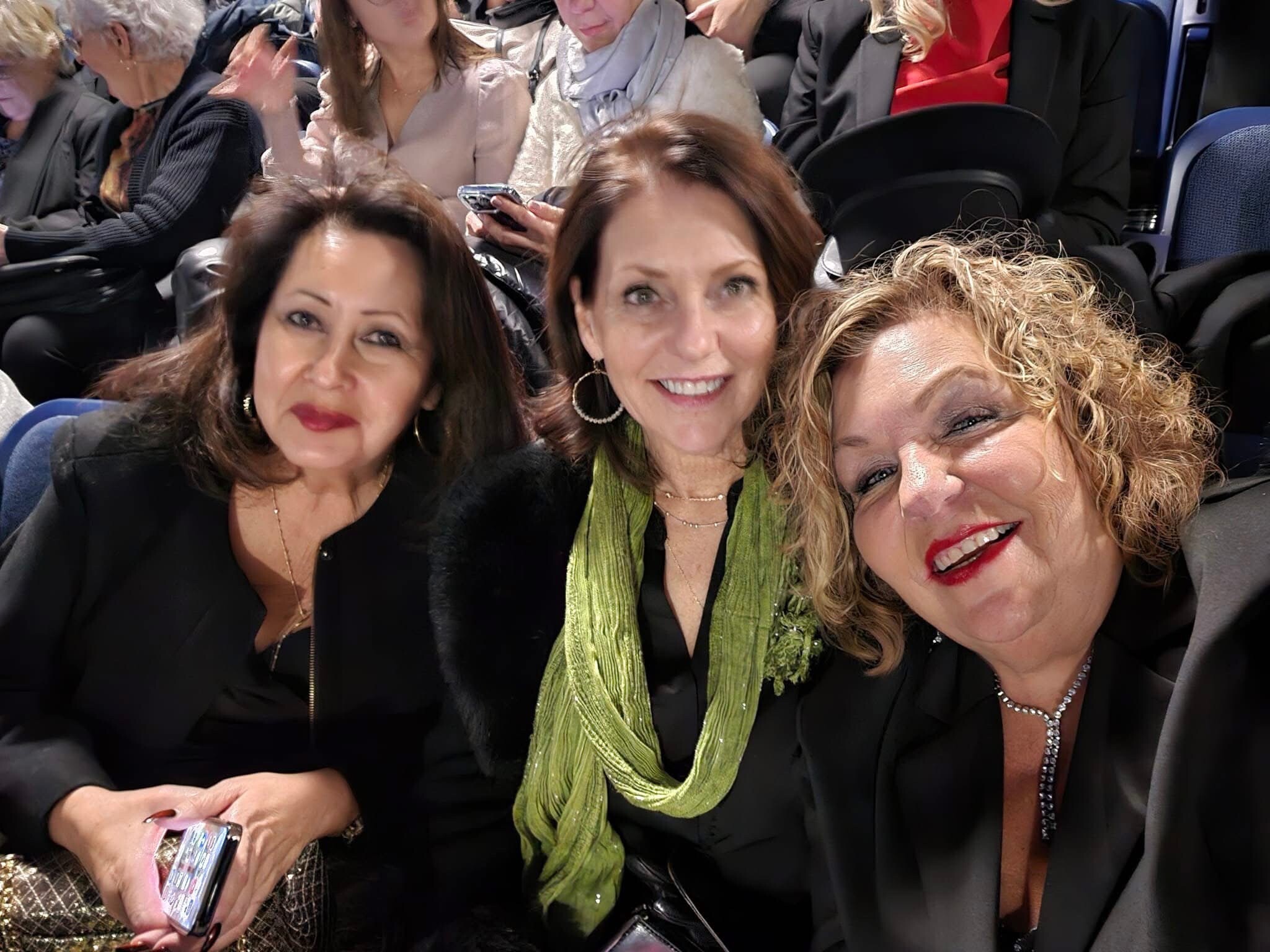 Four women dressed in black, taking a close-up group selfie with smiles, on what appears to be a bus or event setting.
