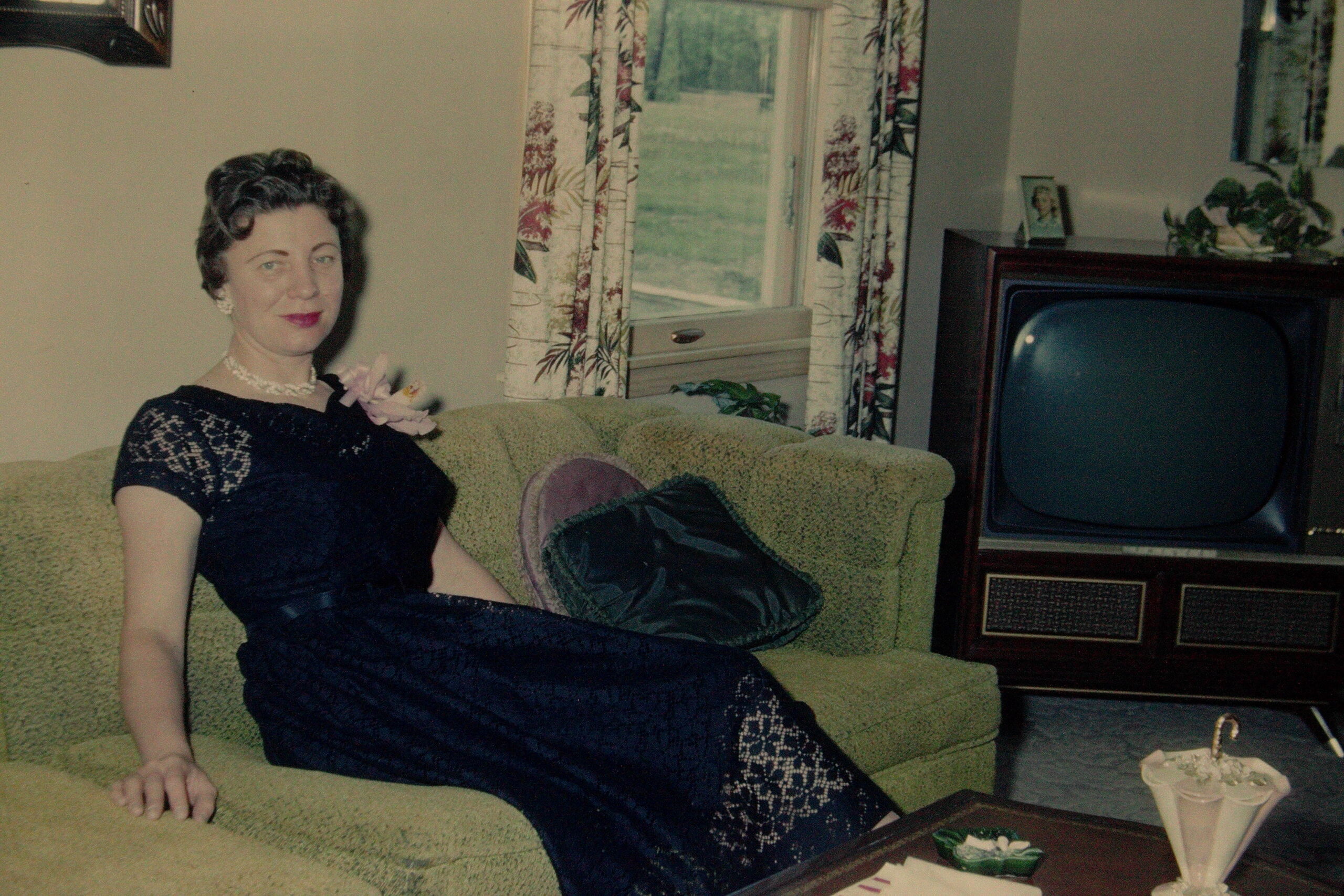 Woman in a dark lace dress sits on a green sofa in a mid-century living room with floral curtains and a vintage television nearby.