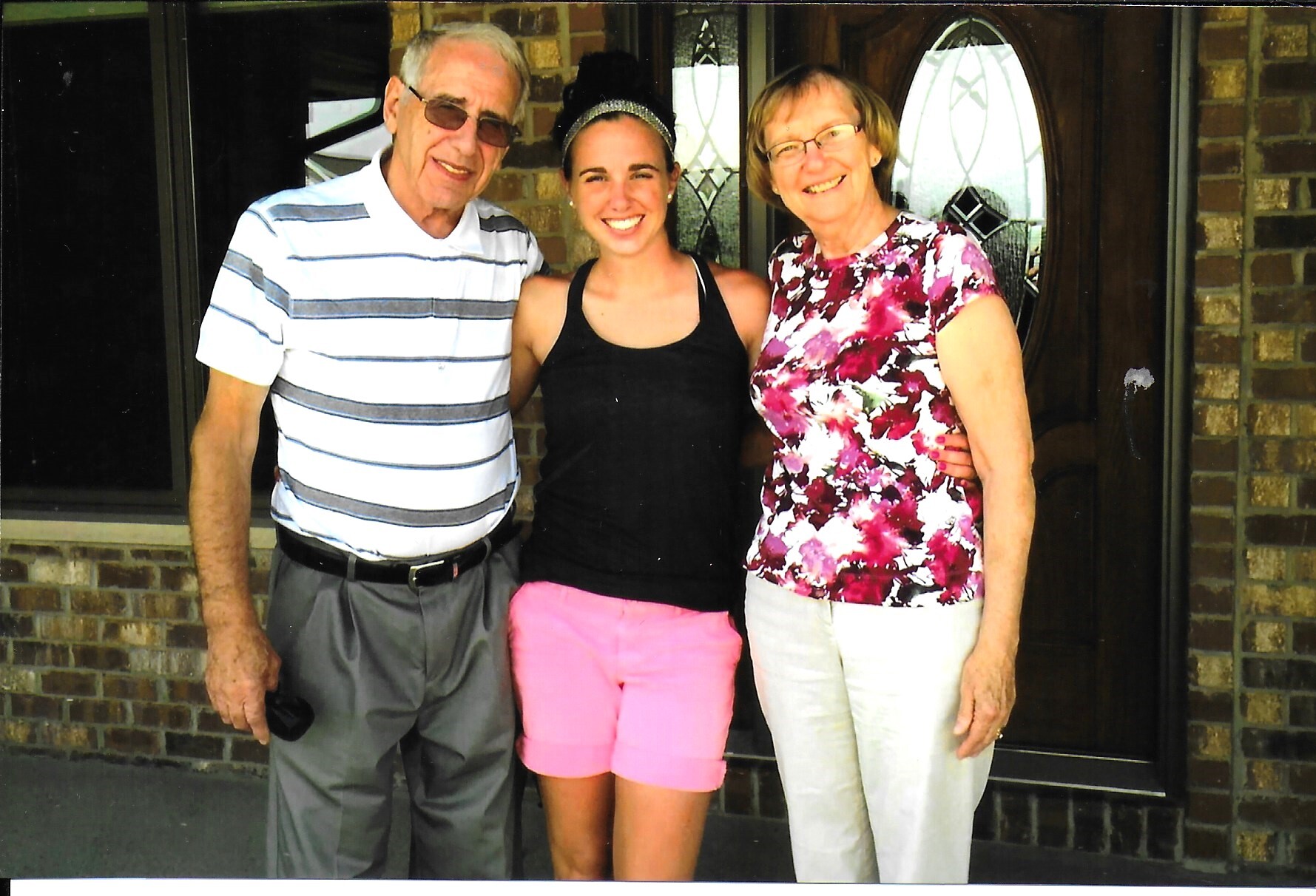 Three people stand arm-in-arm in front of a brick house doorway, smiling at the camera (an older man on the left, a young woman center, an older woman on the right).