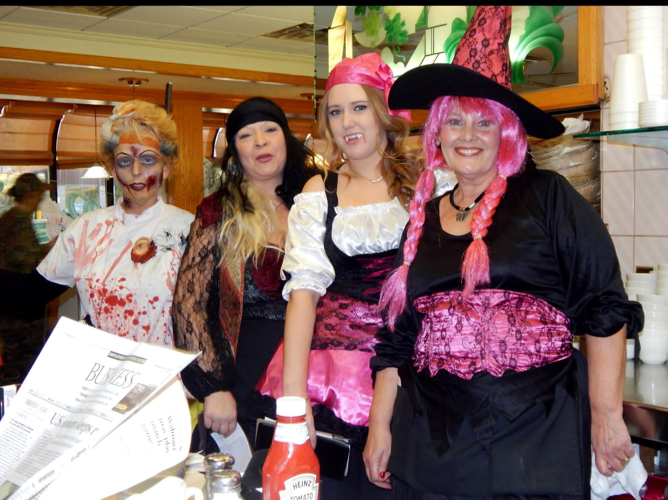 Four women in colorful Halloween costumes pose together for a photo at a party, smiling, with one in pirate attire and another in a zombie-inspired outfit.
