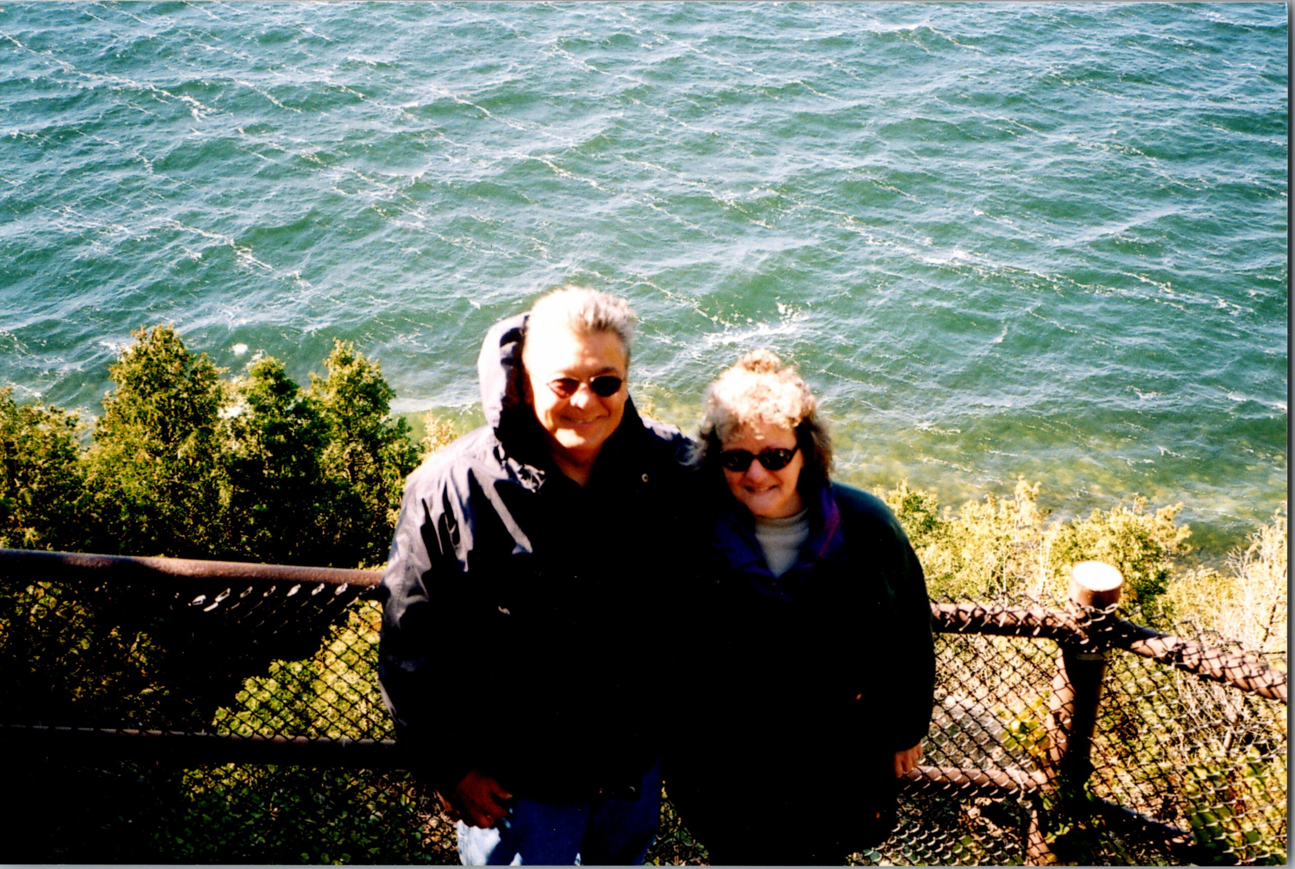 Two adults posing and smiling near a railing with the sea and greenery behind them.