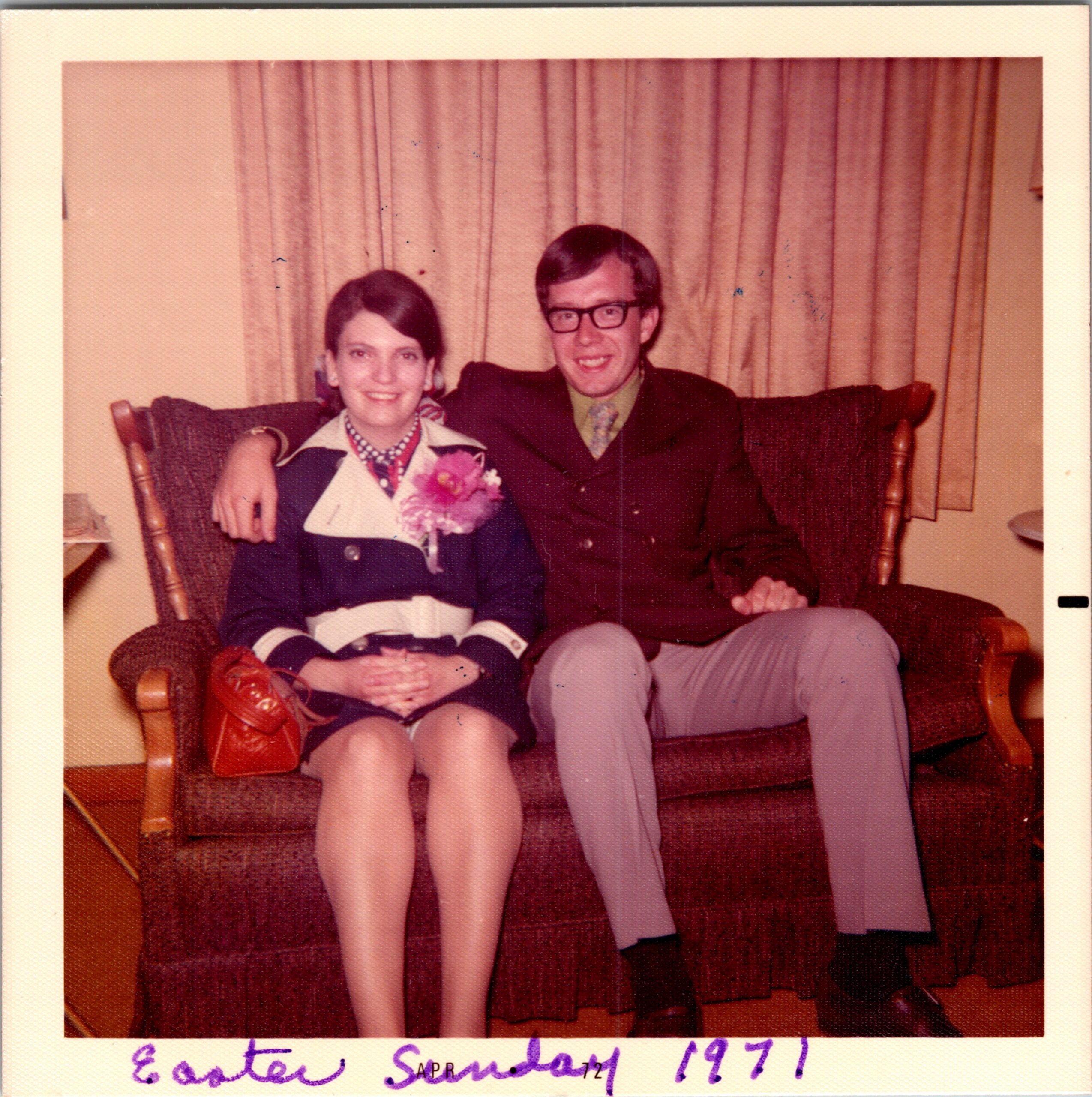 A couple sits together on a brown sofa in a living room; the woman on the left wears a blazer with a corsage, and the man on the right wears a dark suit and glasses, with cream curtains in the background.