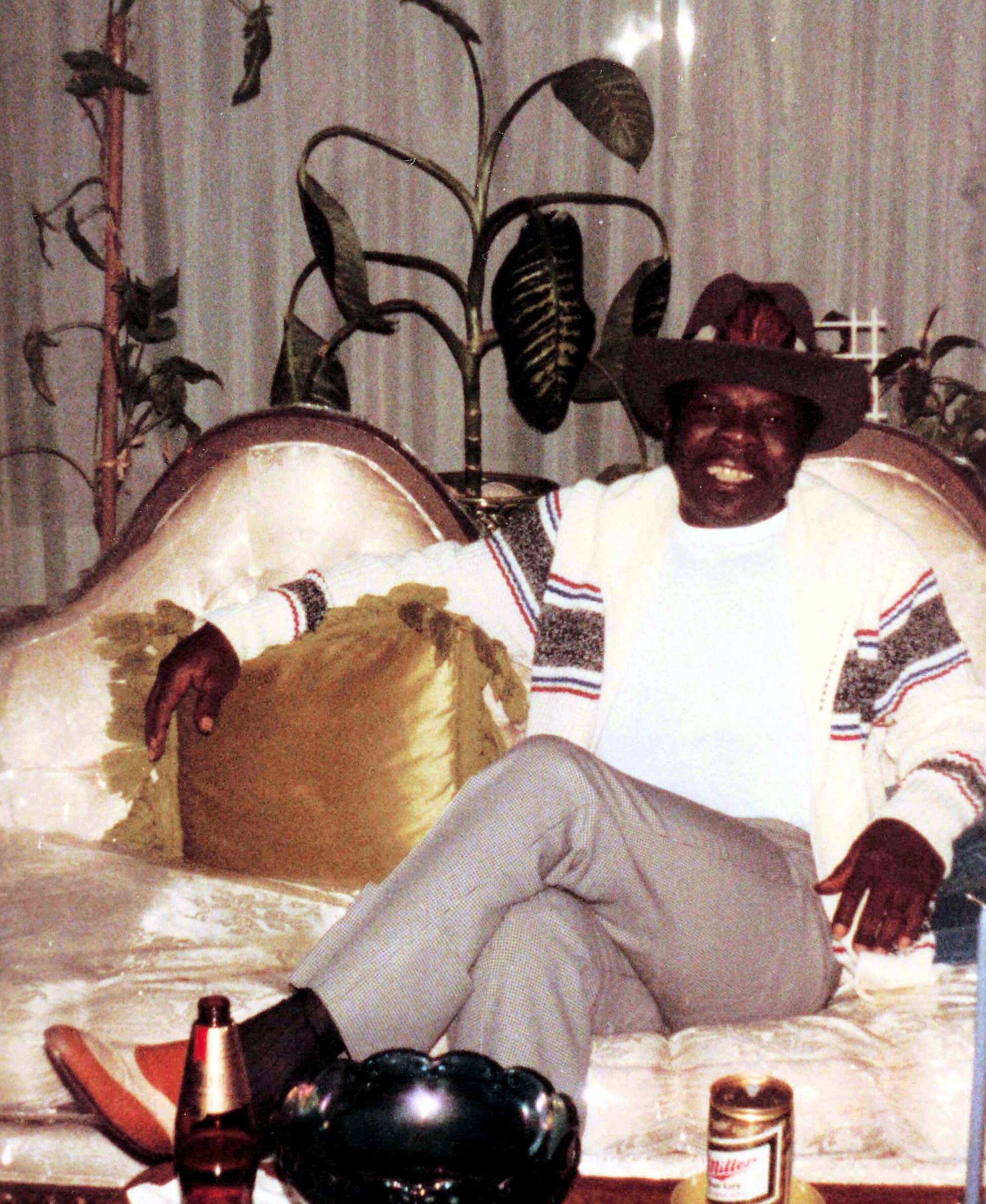 Smiling man wearing a wide-brim hat and a striped poncho seated at a cluttered party table with drinks and a plant in the background