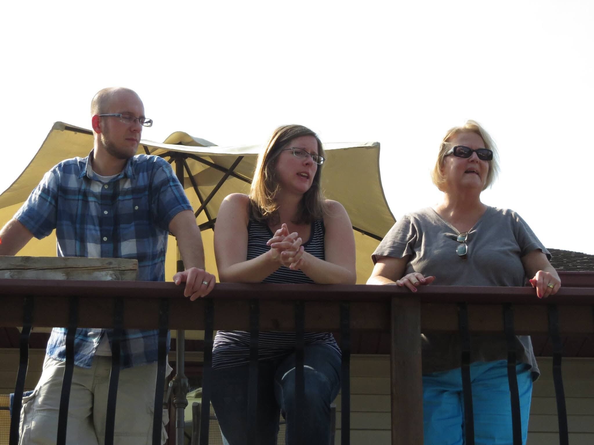 Three adults leaning on a wooden railing on a sunny outdoor deck; left man in a blue plaid shirt, center woman in a striped tank, right woman with sunglasses.