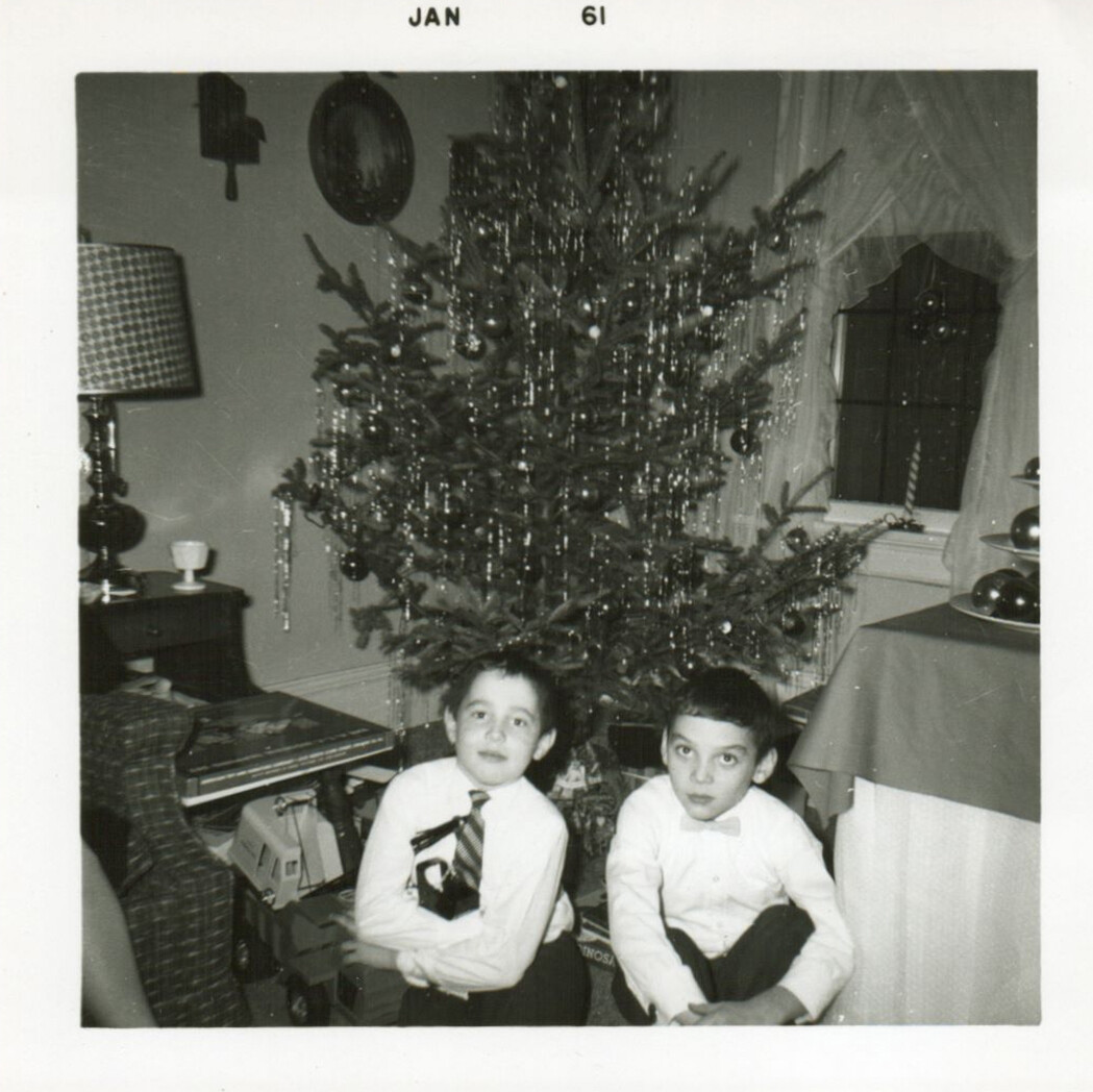 Two young boys in white shirts sit on the floor beside a decorated Christmas tree with presents underneath, in a living room with a piano and lamp to the left and a curtained window to the right.