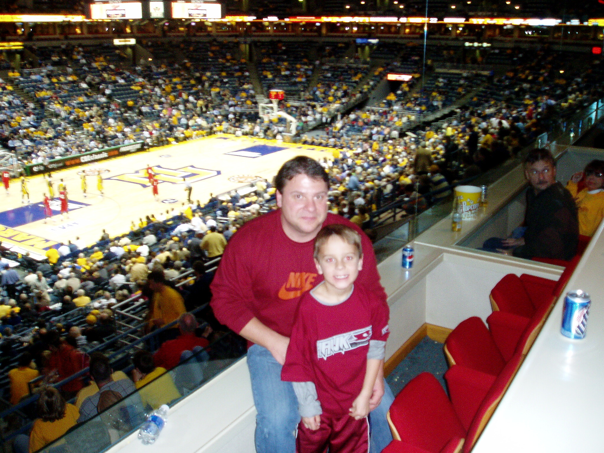 Two boys in maroon shirts pose at a basketball arena railing as a full crowd fills the stands and the court maroons below them with players visible downcourt.