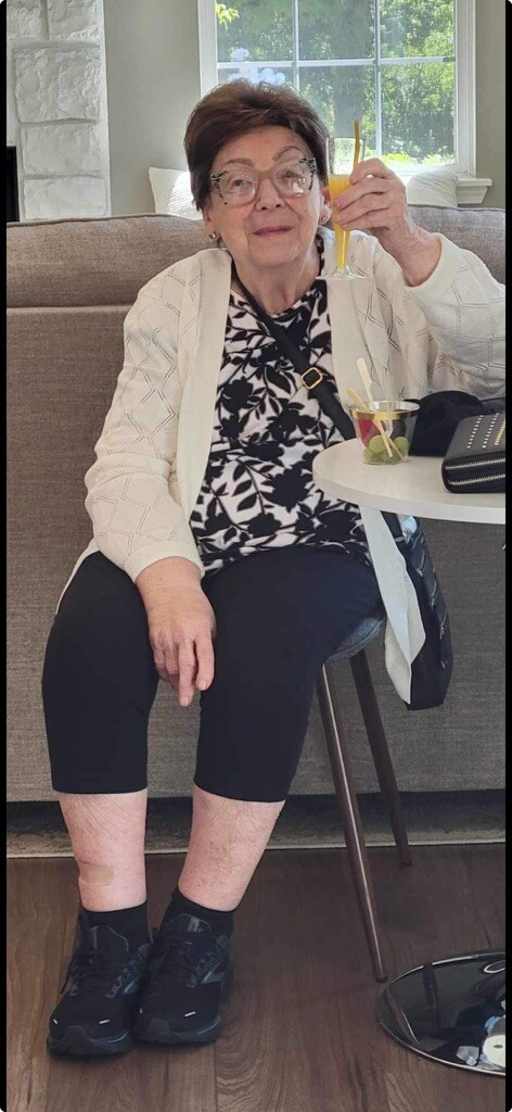 Older woman with short hair and glasses sits on a sofa, raising an orange juice with a yellow straw; a drink and a purse sit on a small table beside her.