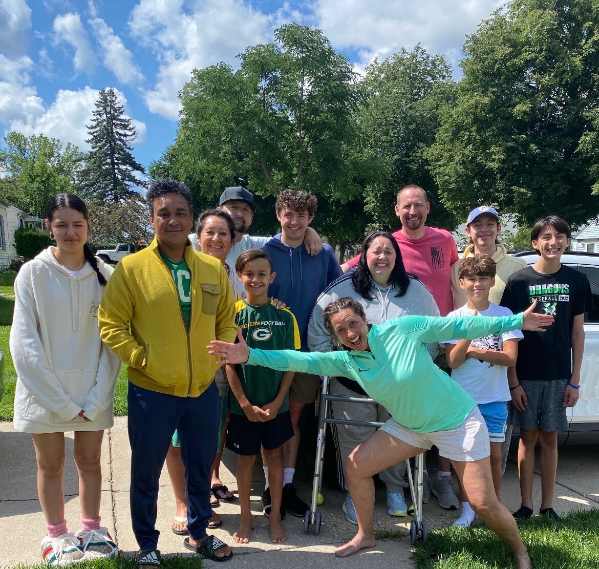 Group of diverse adults and kids posing for a sunny outdoor photo on a residential street; a woman in teal stretches toward the camera in the foreground.