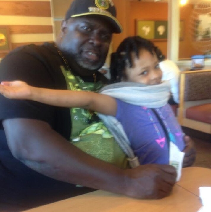 Man wearing a cap sits at a table with a young girl in a purple shirt, both smiling as they pose together in a casual indoor setting.