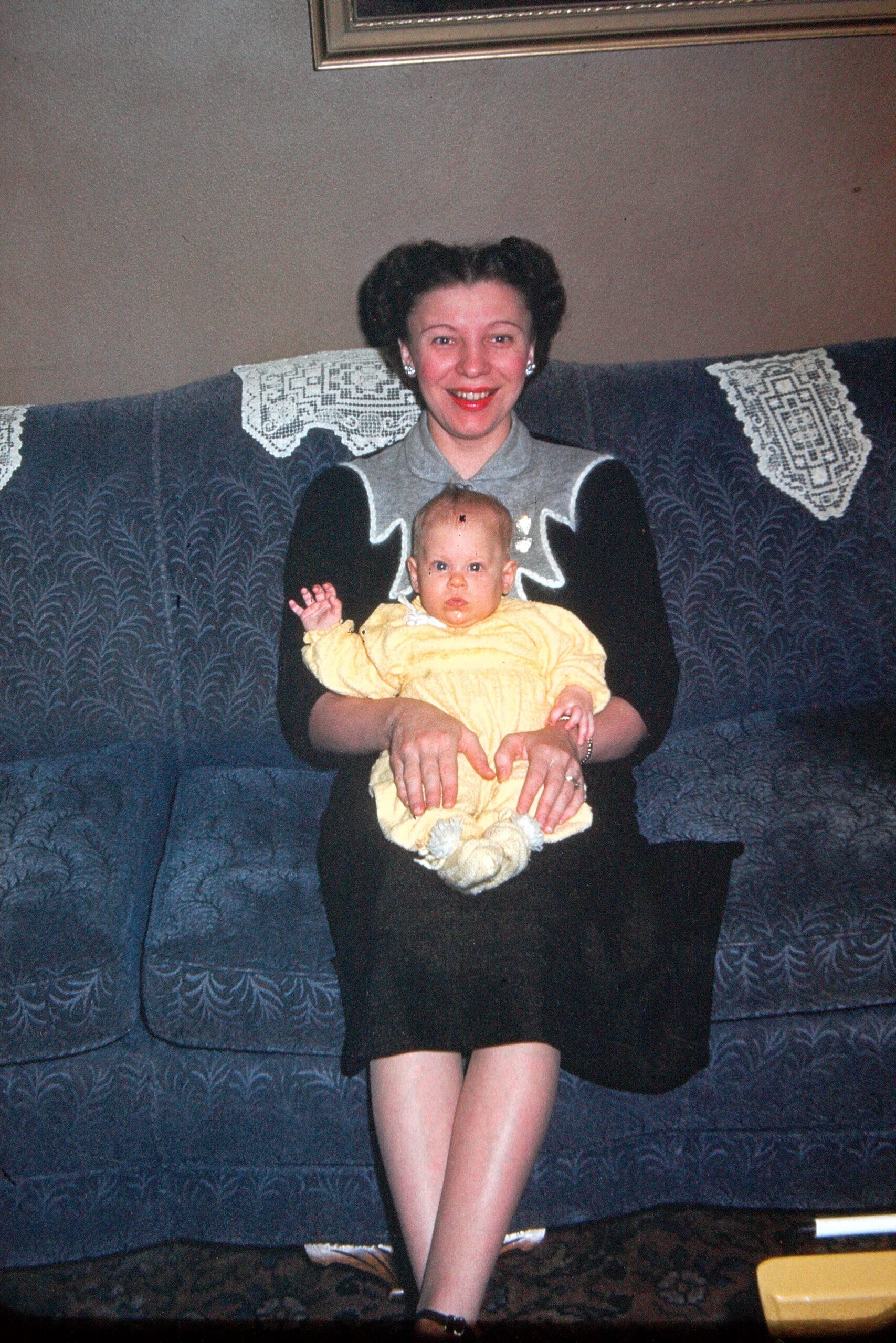 Woman in a gray sweater sitting on a blue patterned couch, holding a baby in a yellow outfit.