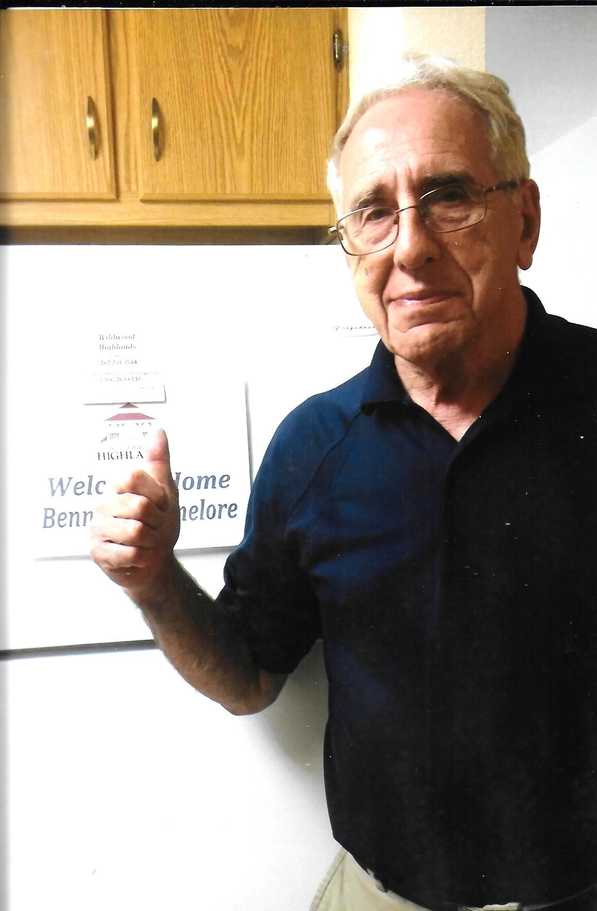 Older man with glasses giving a thumbs-up in a kitchen beside a welcome sign that reads 'Welcome' (partial text visible).