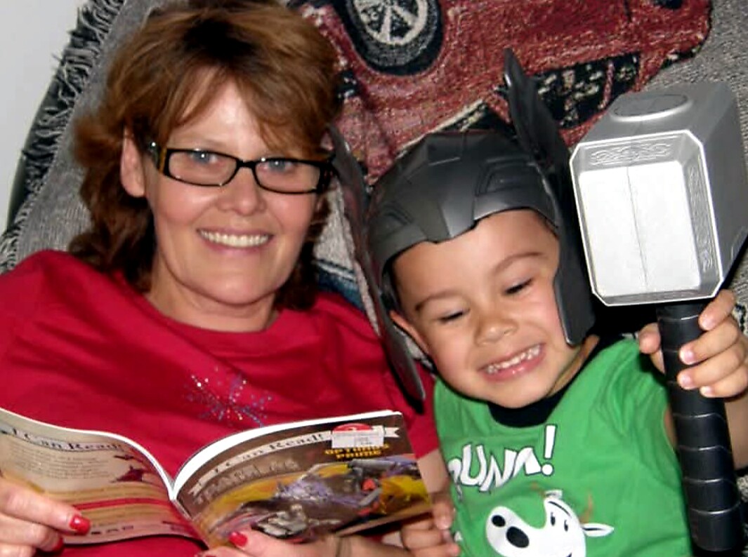 Woman with glasses reading a magazine while a smiling toddler wearing a Batman helmet holds a large toy hammer beside her on the sofa.