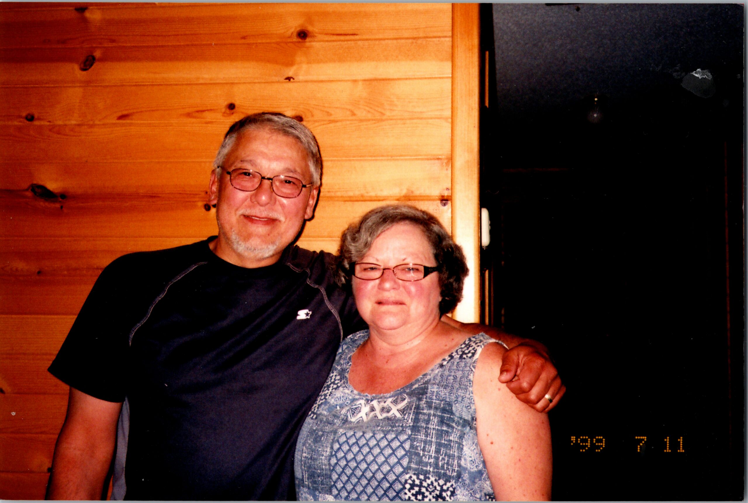 A smiling older couple posing together indoors, man with gray hair and glasses wearing a dark shirt, arm around a woman in a blue patterned sleeveless top, wood-paneled wall in the background.
