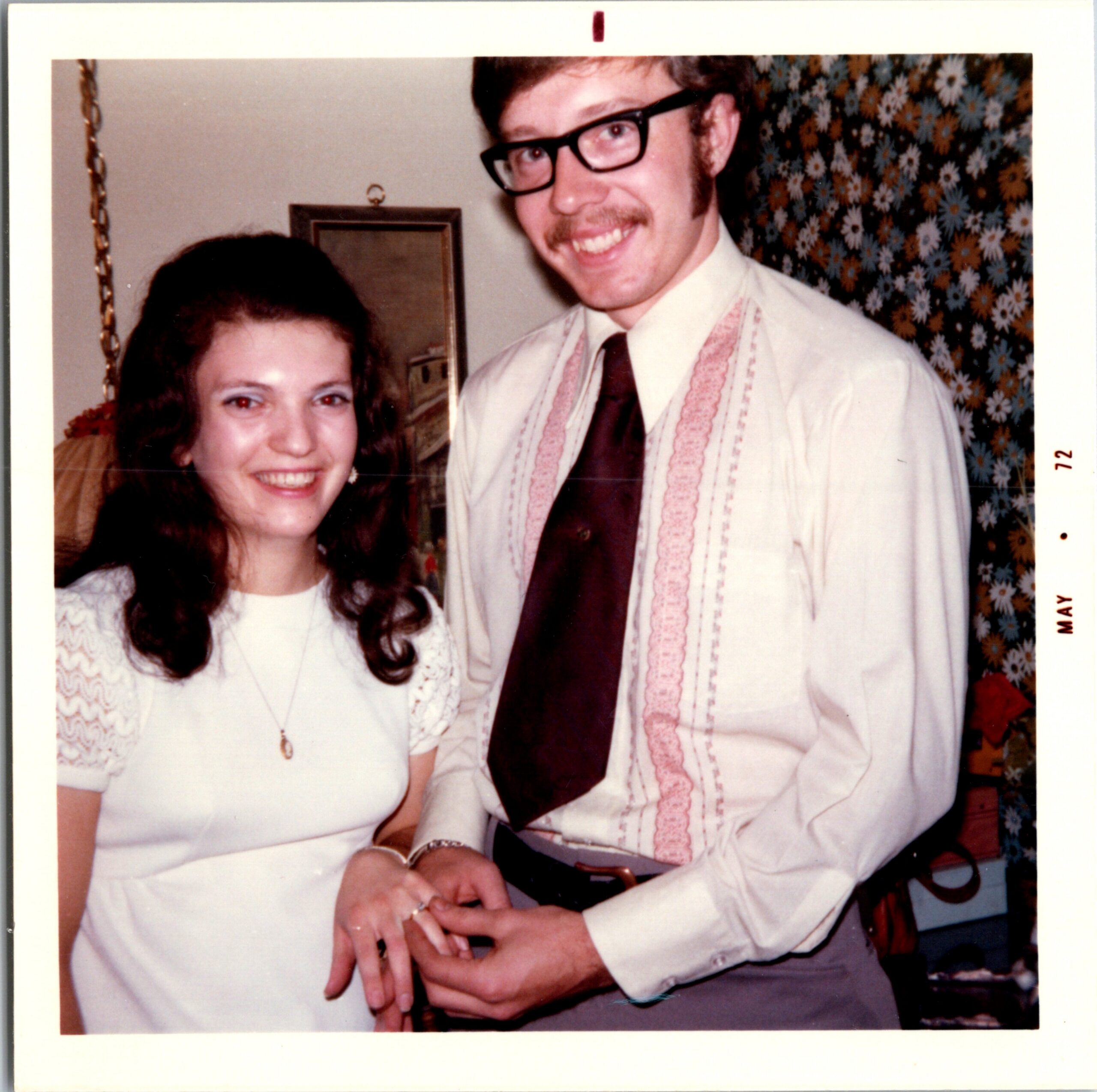 Smiling man in large black glasses and mustache, wearing a light shirt and tie, standing next to a woman in a white lace top; retro floral wallpaper in background.