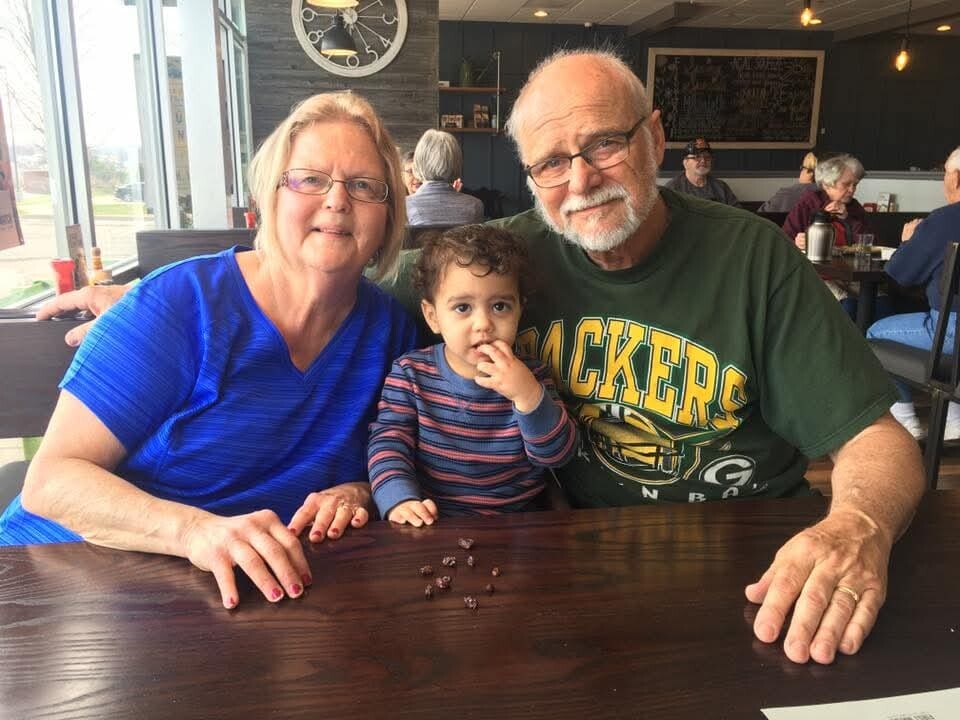 Grandparents and a young child sit together at a restaurant table; the child is nibbling on a chocolate piece.