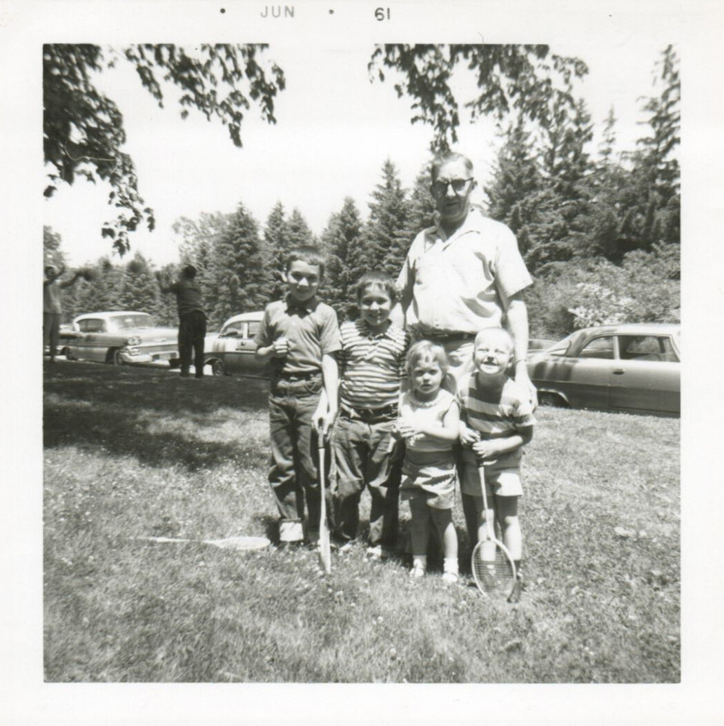 Family group outdoors: an adult man stands with four children on a grassy park, two kids hold tennis rackets, vintage cars in the background (June 1961).