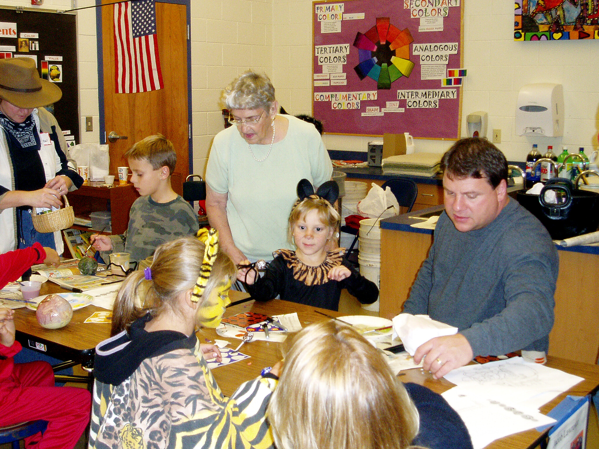 Group of children and adults crafting at a long table in a classroom, with art supplies and costumes visible on the table and walls in the background.