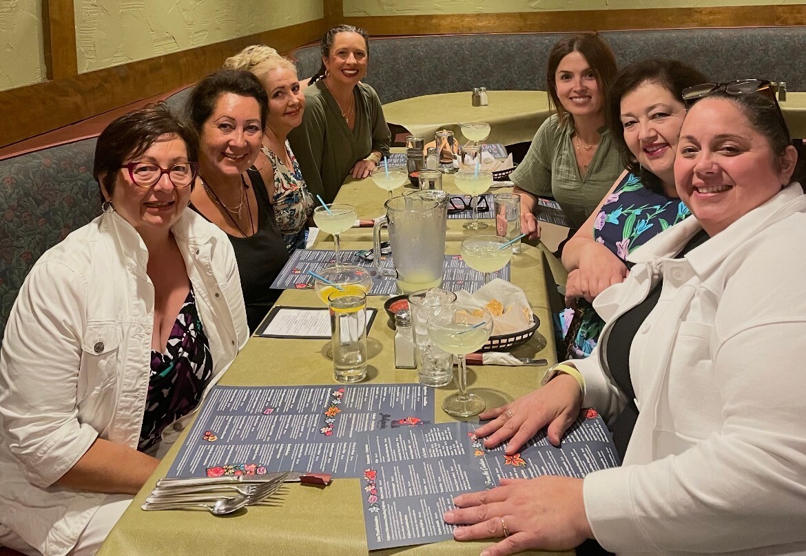 Seven women sit around a restaurant table smiling at the camera during a meal together.