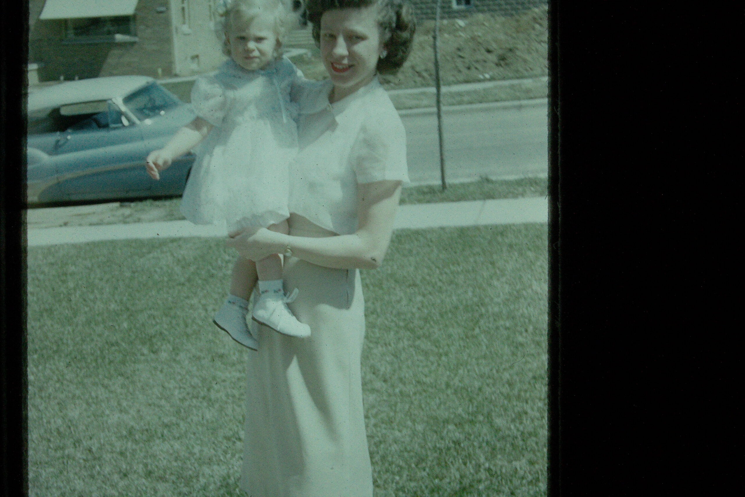 A woman in a light dress holds a young child outdoors, with a vintage car in the background.