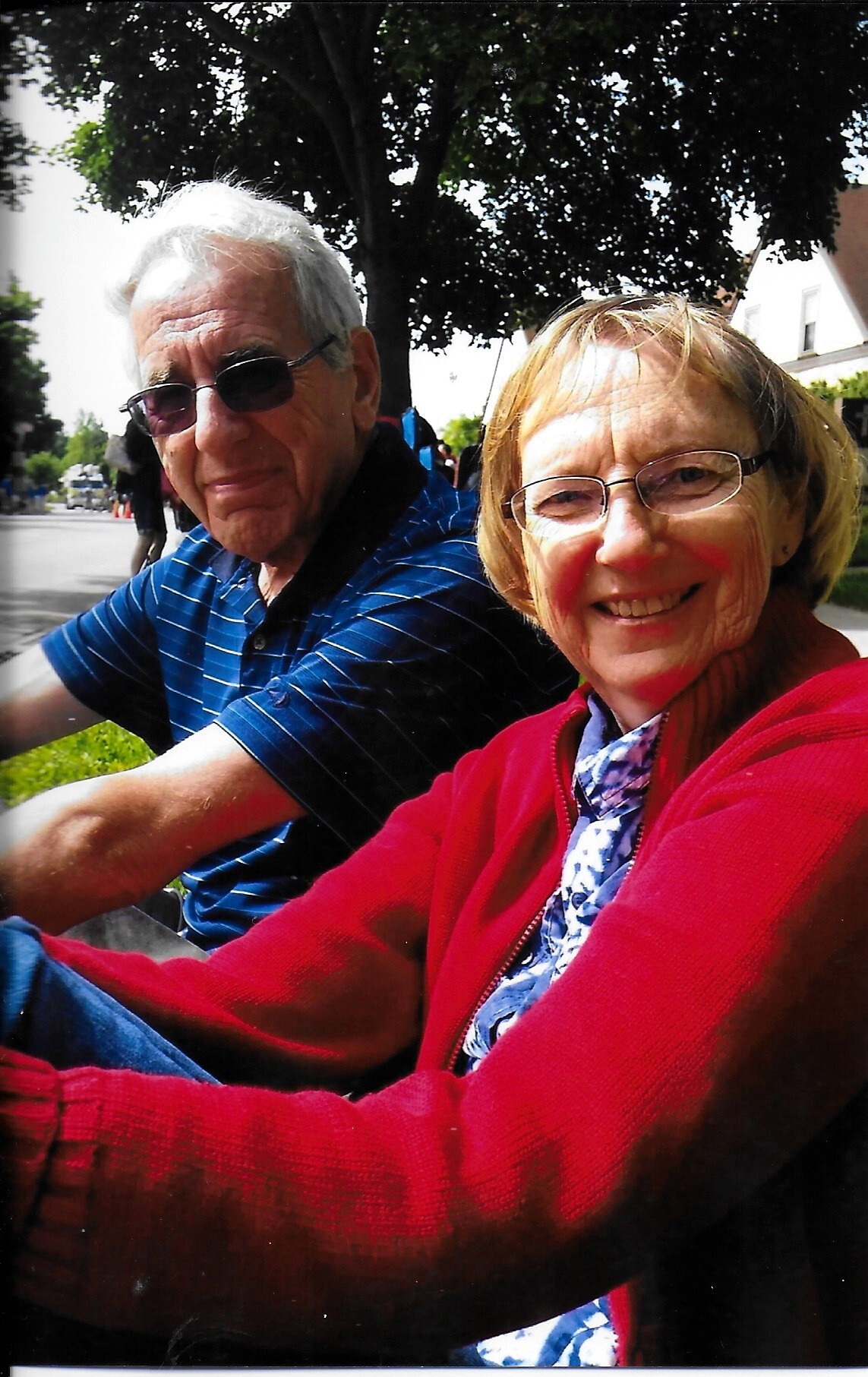 Smiling elderly couple posing for a selfie outdoors, woman in a red cardigan and glasses, man wearing sunglasses beside her under a leafy tree.