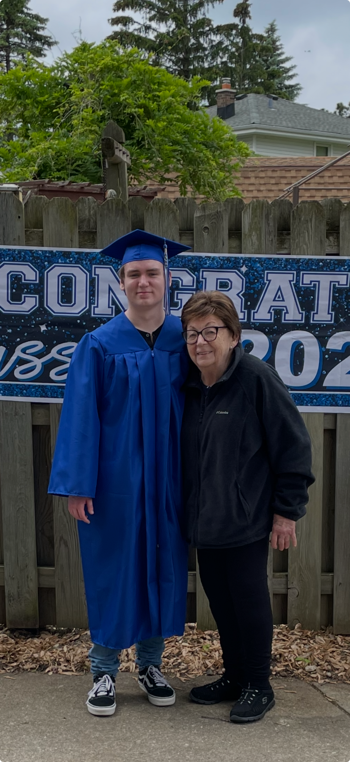 Graduate in a blue cap and gown posing with a woman in a black jacket in front of a large 'Congratulations' banner outdoors.
