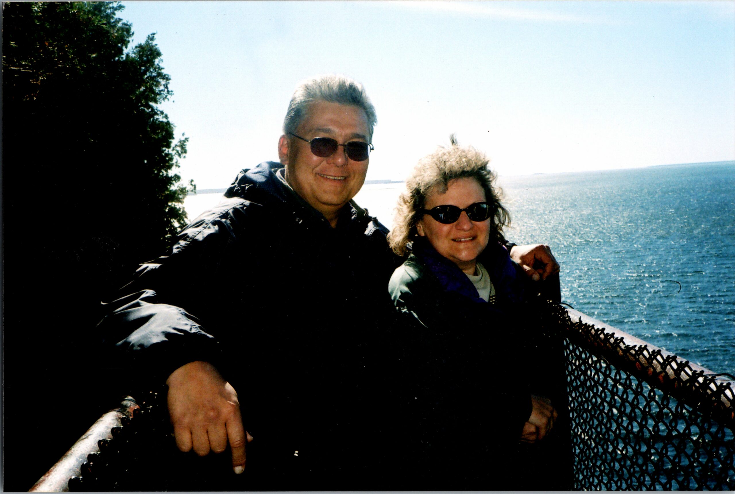 Two people wearing sunglasses lean on a metal railing by a body of water on a sunny day, smiling at the camera.
