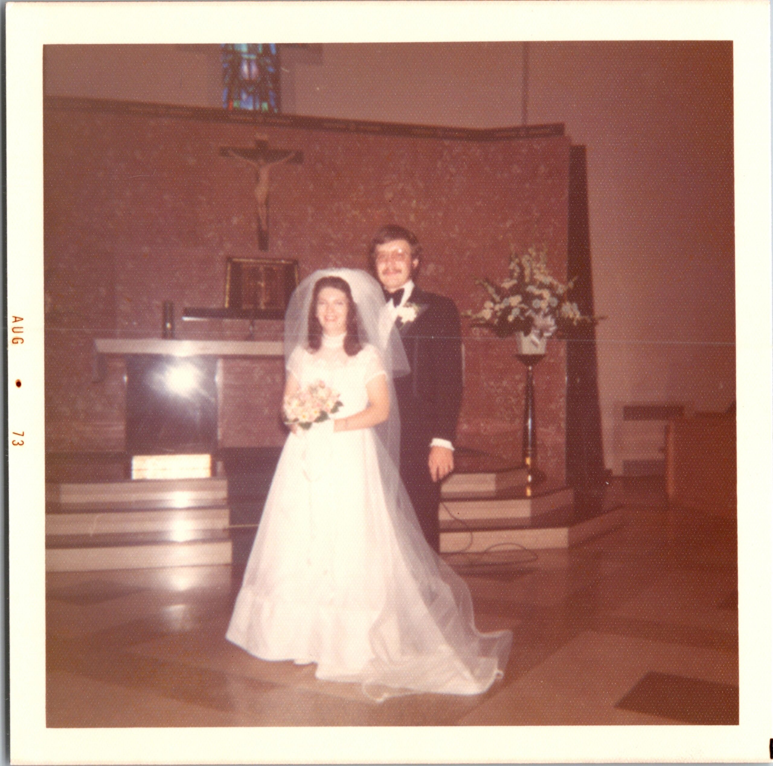 Bride in a white gown and veil holds a bouquet beside the groom in a tuxedo at a church altar with a crucifix and flowers behind them.