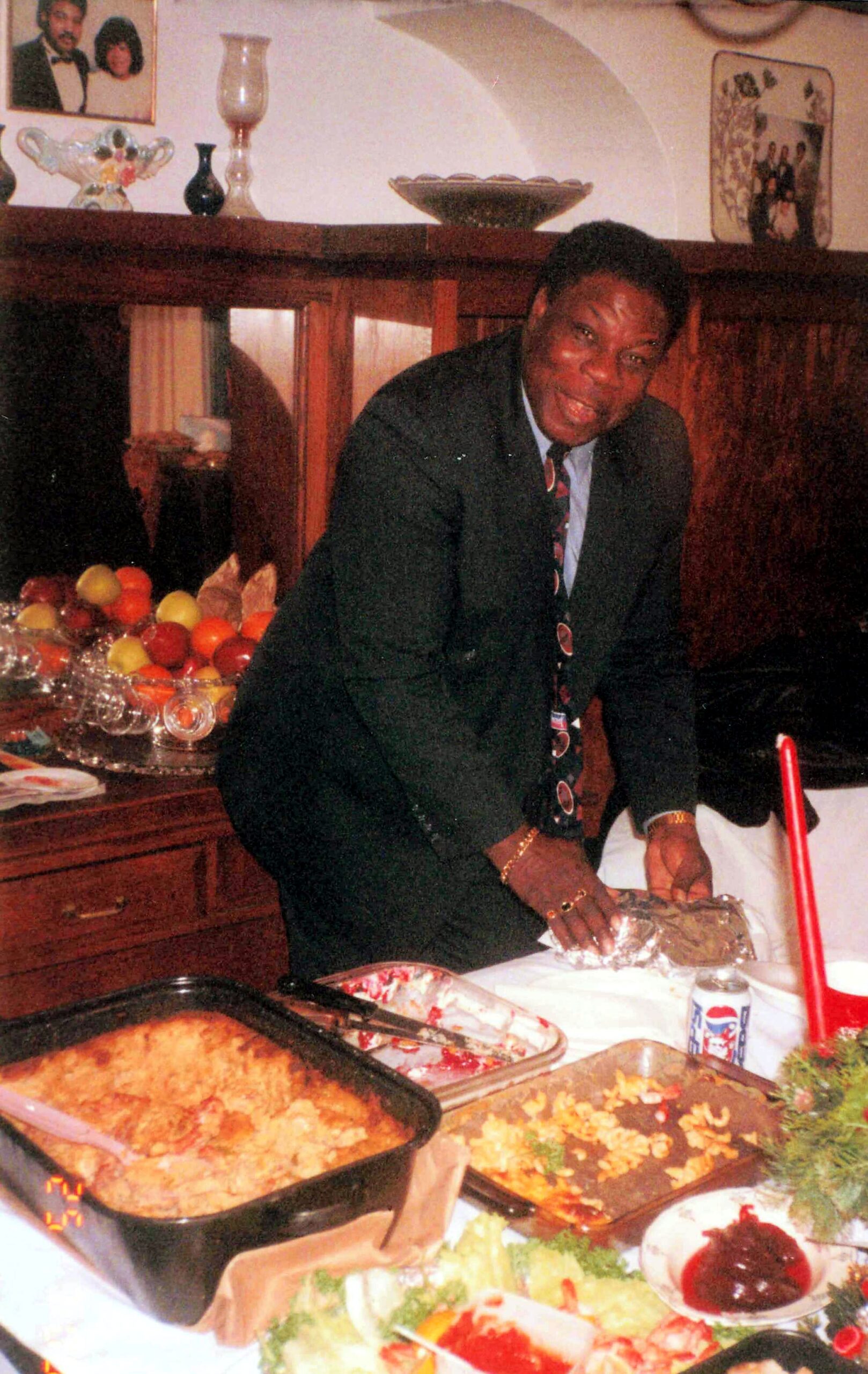 Smiling man in a dark suit and patterned tie leaning over a table of food at a home buffet.