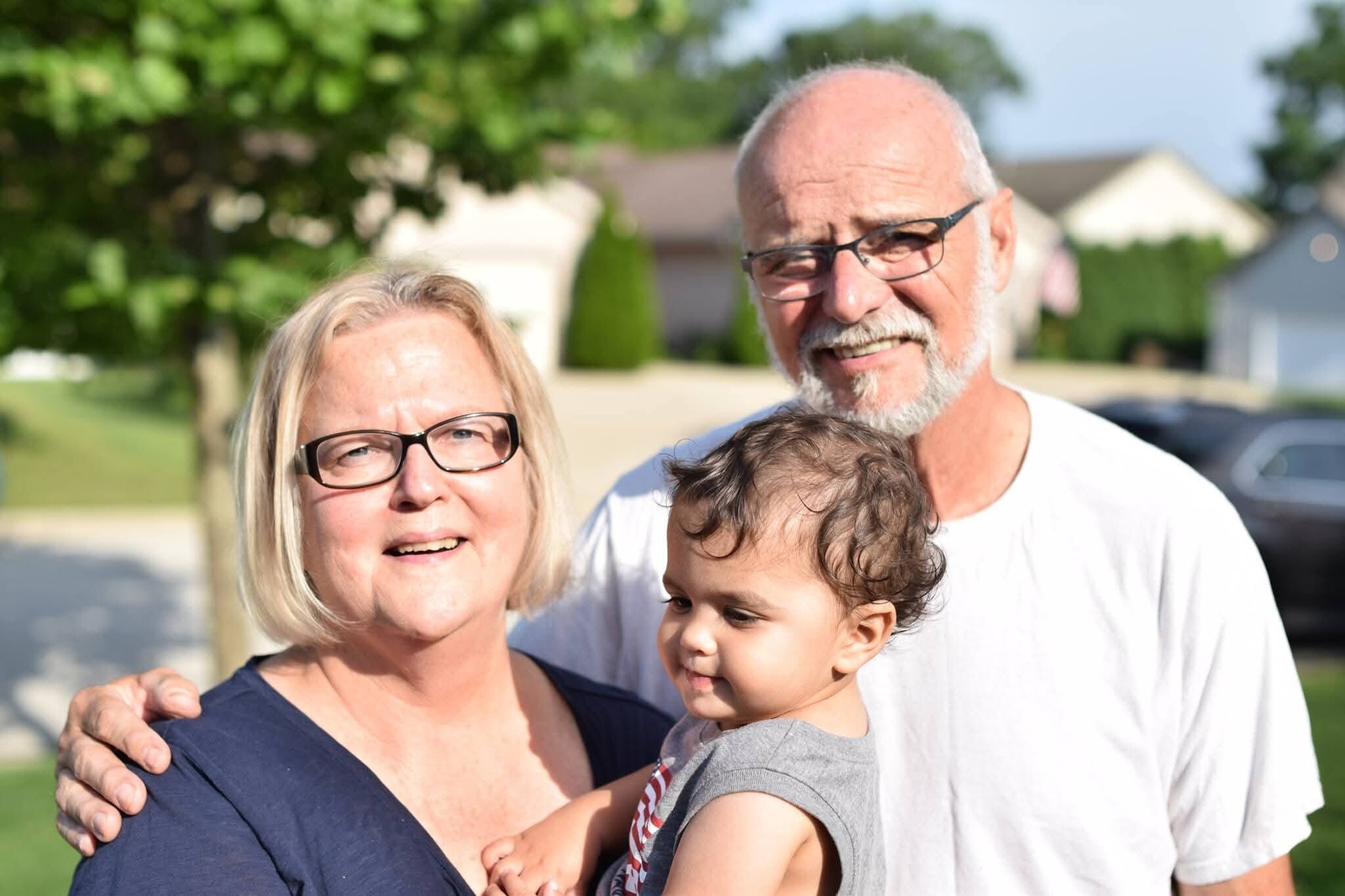 A smiling older couple with glasses posing outside with their young grandchild, in a sunny suburban neighborhood.