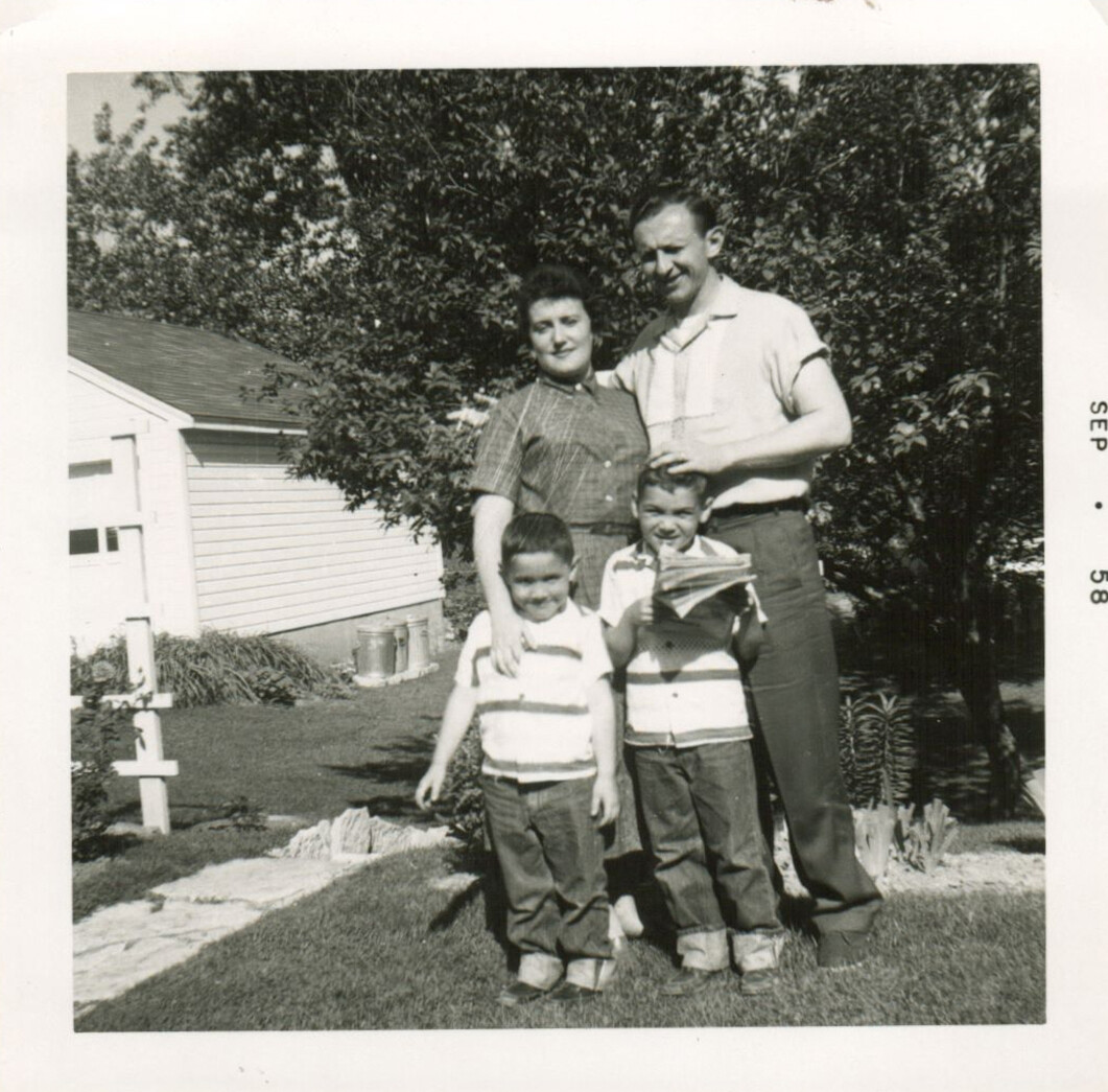 Black-and-white family photo: two adults standing behind two children in a front yard with a house and trees in the background.