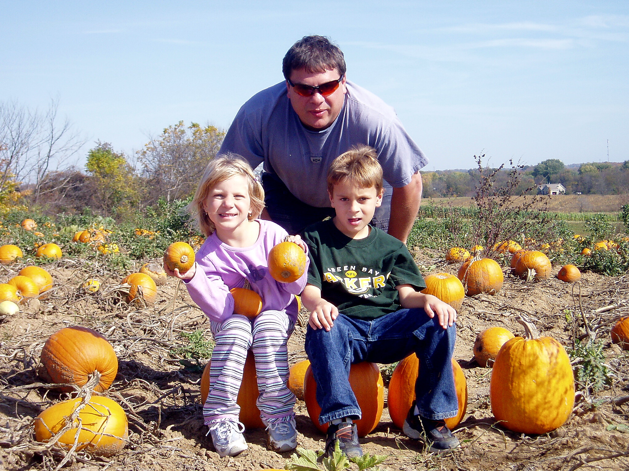 Man and two children posing in a pumpkin patch among ripe orange pumpkins on a sunny day, smiling at the camera.