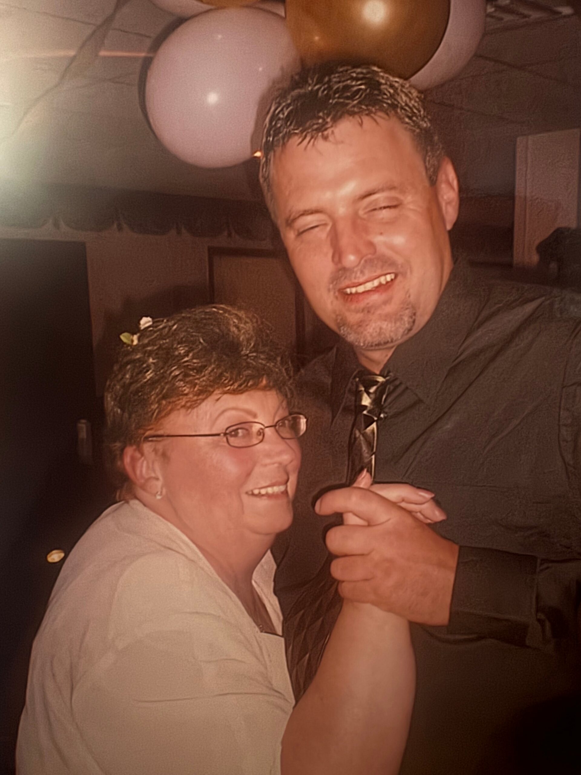 Smiling couple dancing at a party indoors, woman wearing glasses and a light top, man in a dark shirt, balloons overhead.